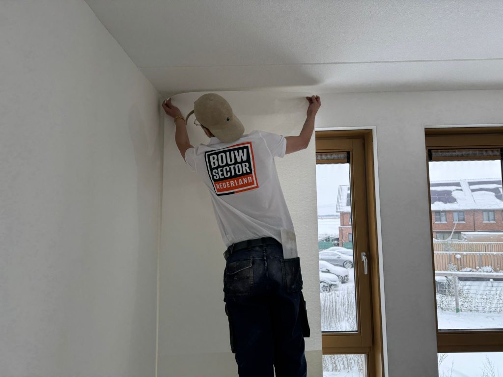 Behanger van Bouwsector Nederland drukt een baan renovlies bovenaan tegen de wand, onder een wit plafond